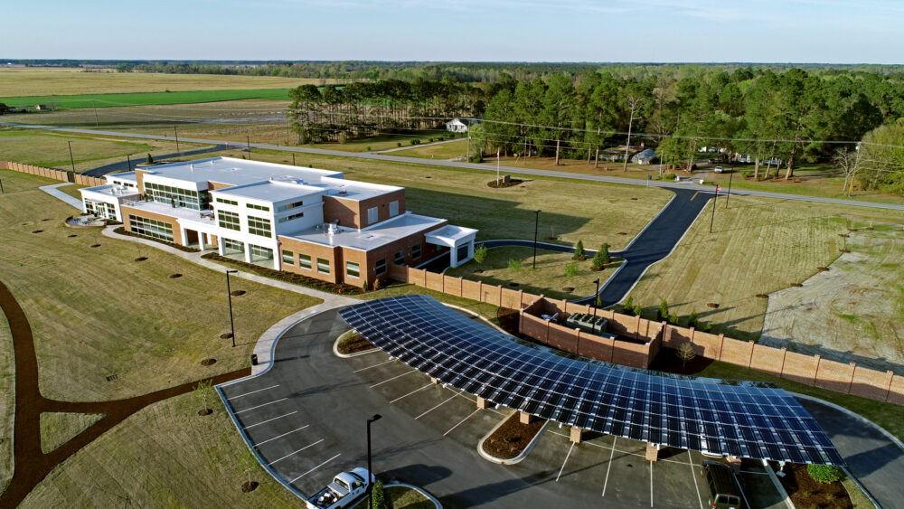 Aerial view of Solar Carport using custom LSX Solar panels built over parking lot surrounded by grass with two cars parked and Marlboro Electric Coop building in background in South Carolina