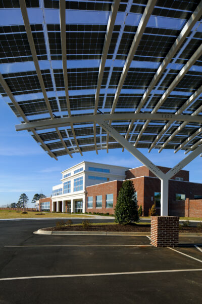 Underside view of Solar Carport using custom LSX Solar panels built over parking lot surrounded by grass with two cars parked and Marlboro Electric Coop building in background in South Carolina