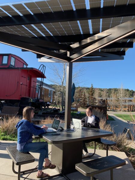 SolarZone Solar Charging Work Table on outside patio in front of train car with two people working on laptops in public space with trees in background on a sunny day