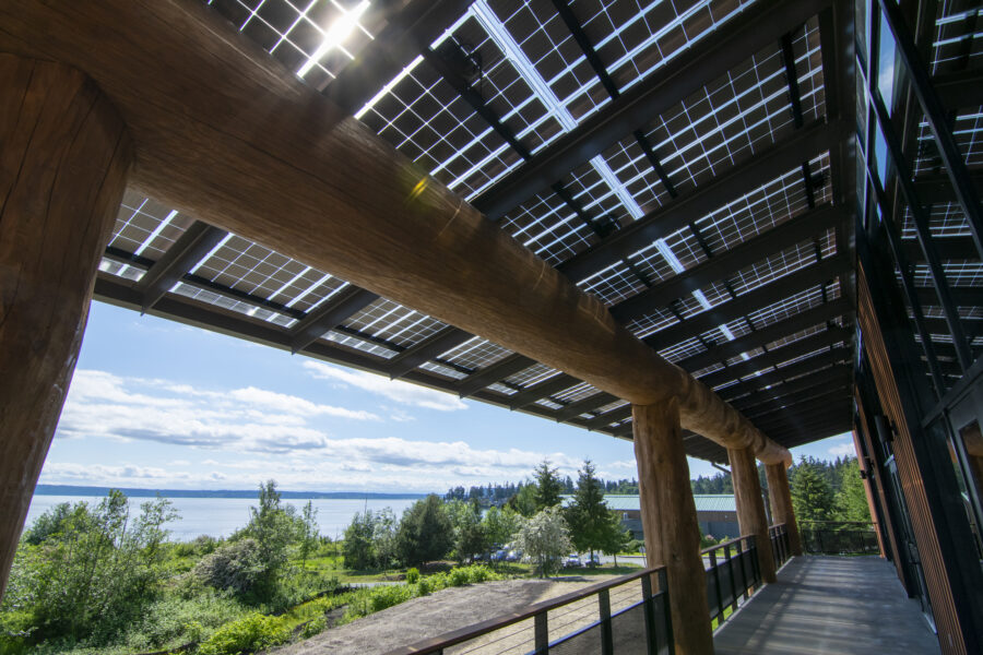 LSX Solar Panel Awning covering wraparound deck outside of Tulalip Gathering Hall with large wooden beams, looking out at view of lake and trees in background
