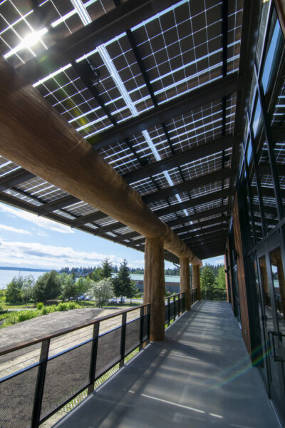 LSX Solar Panel Awning covering wraparound deck outside of Tulalip Gathering Hall with large wooden beams and view of lake in background.