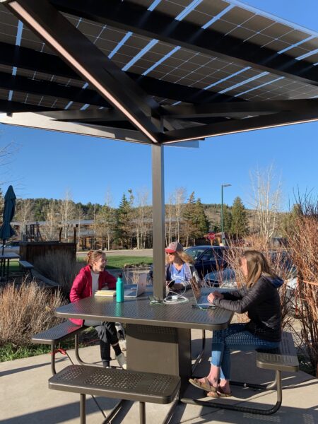 Solar Charging Table in town center in front of a restaurant with people sitting at benches and talking while working on computers and charging devices.