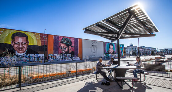 Solar Charging Table in commercial outdoor space in front of a wall mural with people sitting at benches and working on laptop computers. PIA 2020 Product Innovation Award Winner.