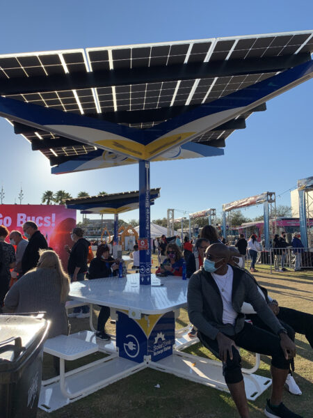 Solarzone Recharge Station with people sitting on benches under solar shade and charging phone devices.