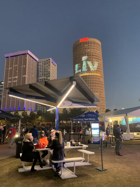 Solarzone Recharge Station at NFL Super Bowl LV Experience at night time with people sitting on benches under solar shade with LED Lighting and charging phone devices.